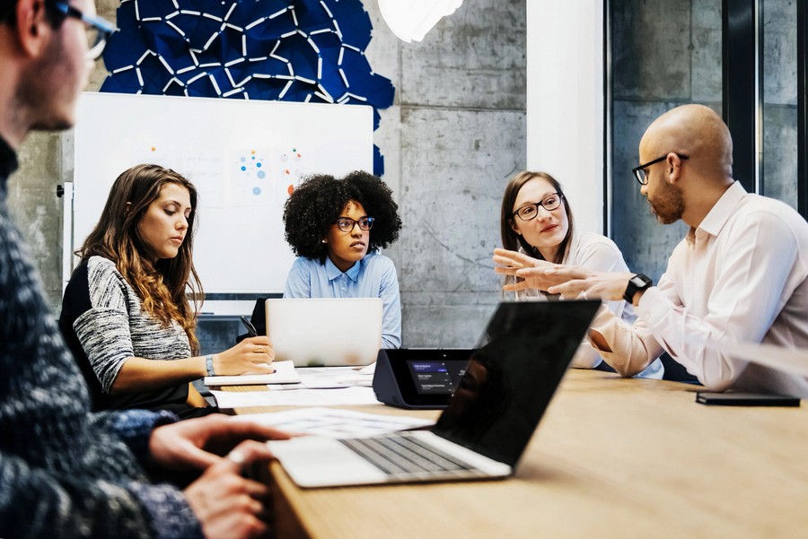 Five professionals sit at a conference table with laptops, discussing ideas during a business meeting.